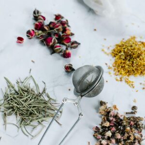 A flat lay of herbal tea ingredients with a tea strainer on a marble surface.